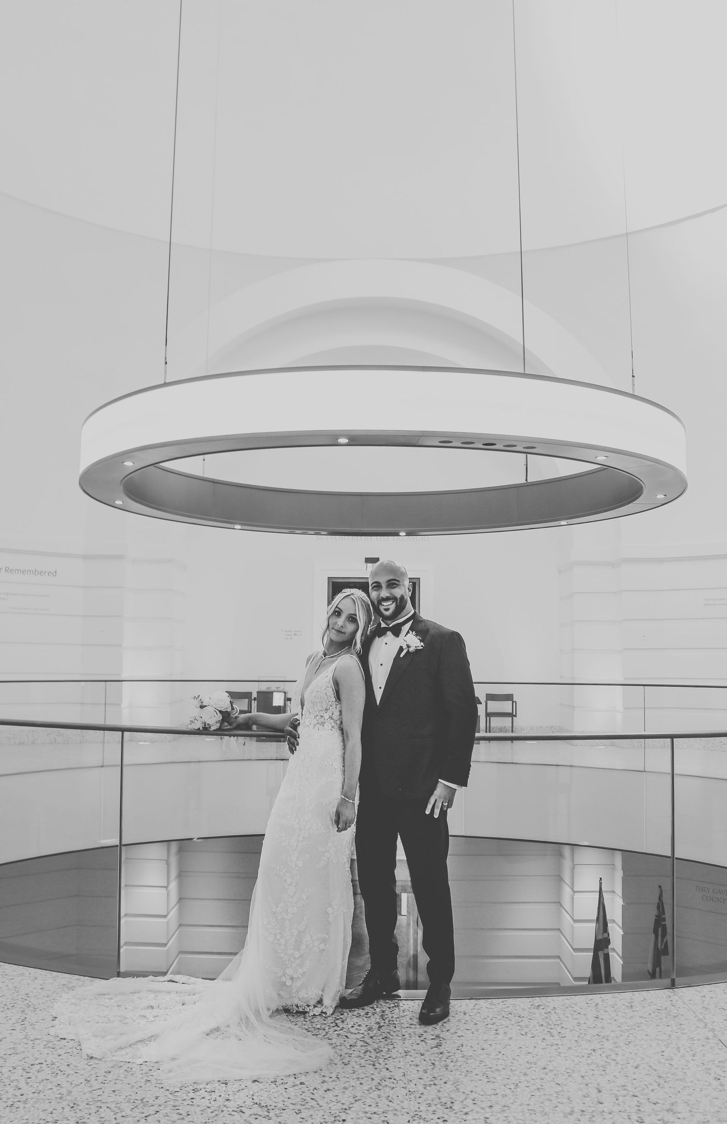 A black and white photograph of newlyweds posing under a modern circular light fixture in a contemporary space.