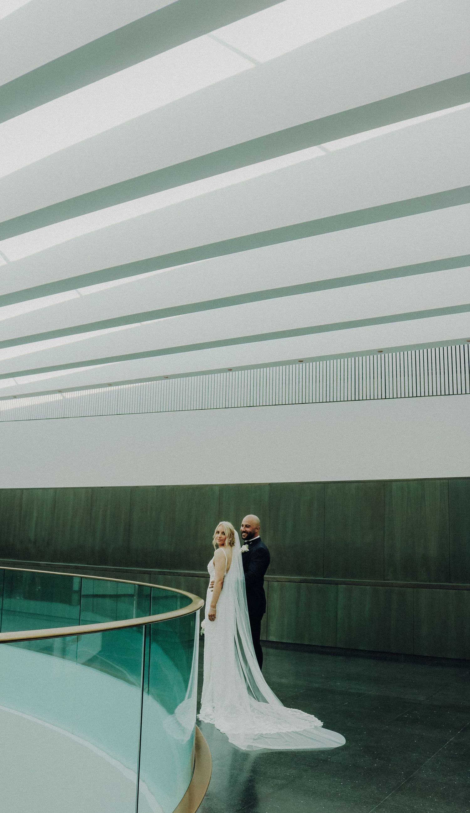 A newlywed couple stands in a modern architectural space with curved railings, mint green walls and geometric ceiling details.