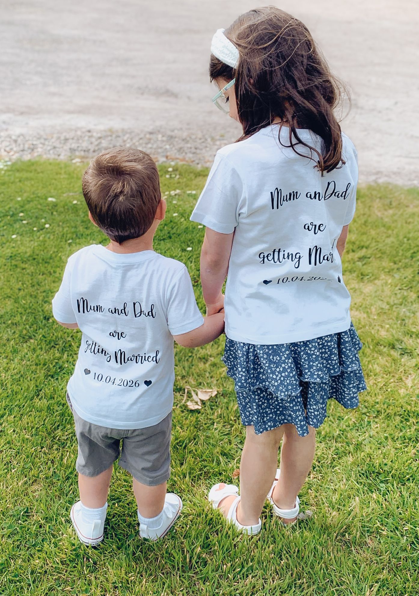 Two kids wearing matching white t-shirts with text about mom and dad getting married standing on grass outdoors.