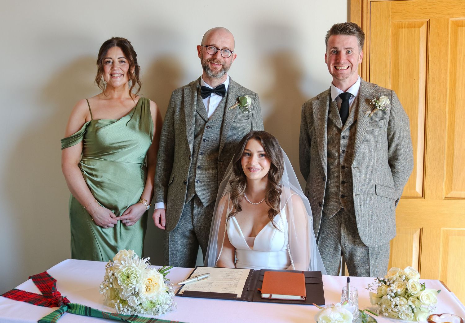 A wedding party stands behind a table with white floral arrangements and a signing book during a civil ceremony.