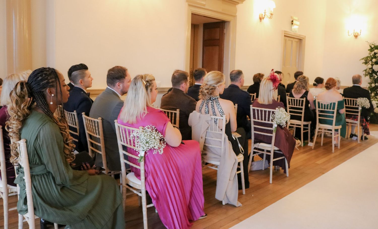 Wedding guests seated in white Chiavari chairs line the walls of an elegant indoor venue during a ceremony.