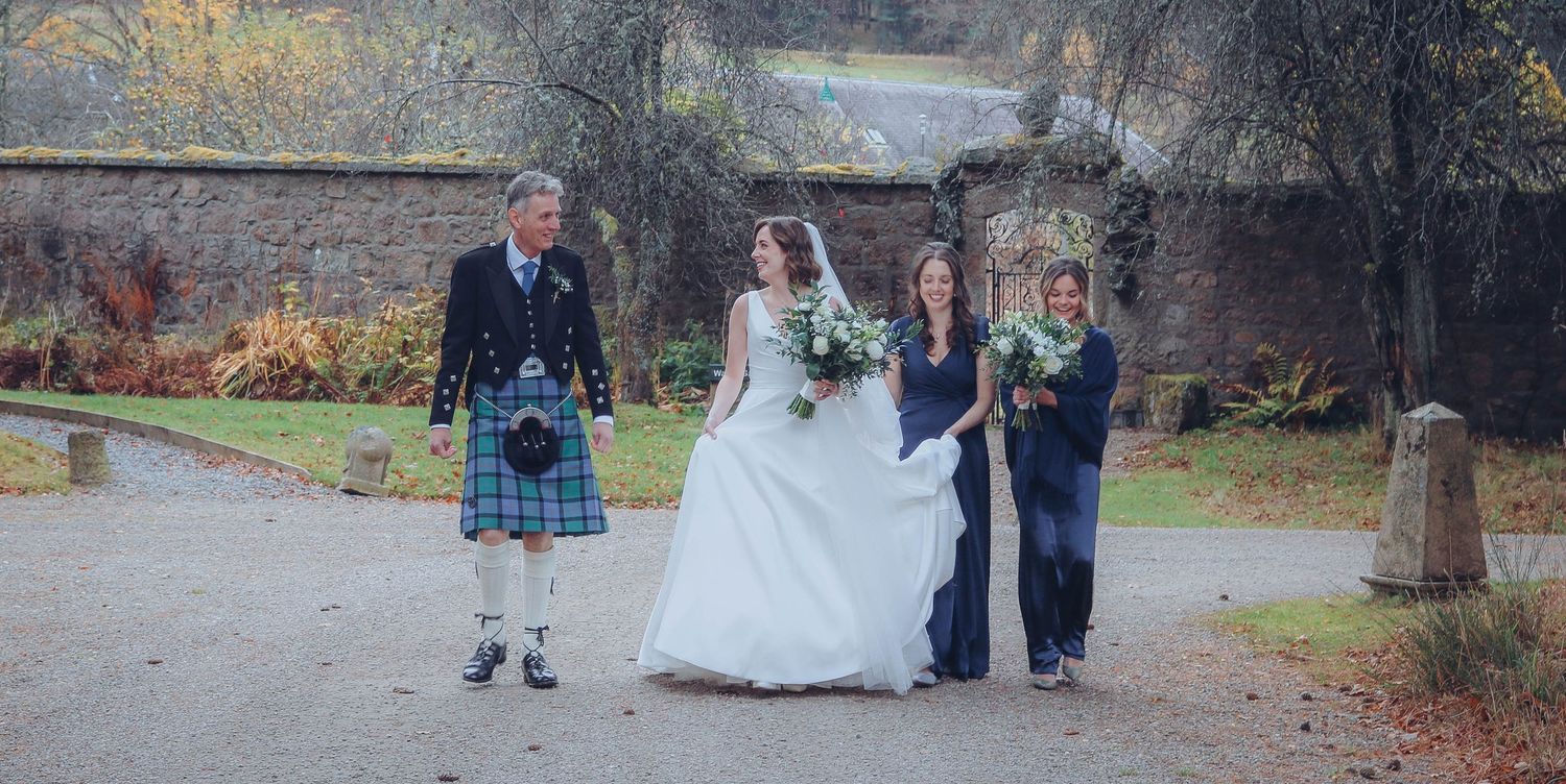 Four people in formal attire including traditional Scottish kilt and white wedding dress walk together on garden path.