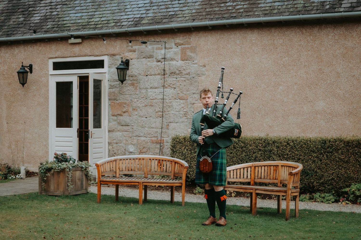 Traditional bagpiper in green kilt performs in garden with wooden benches outside stone building.