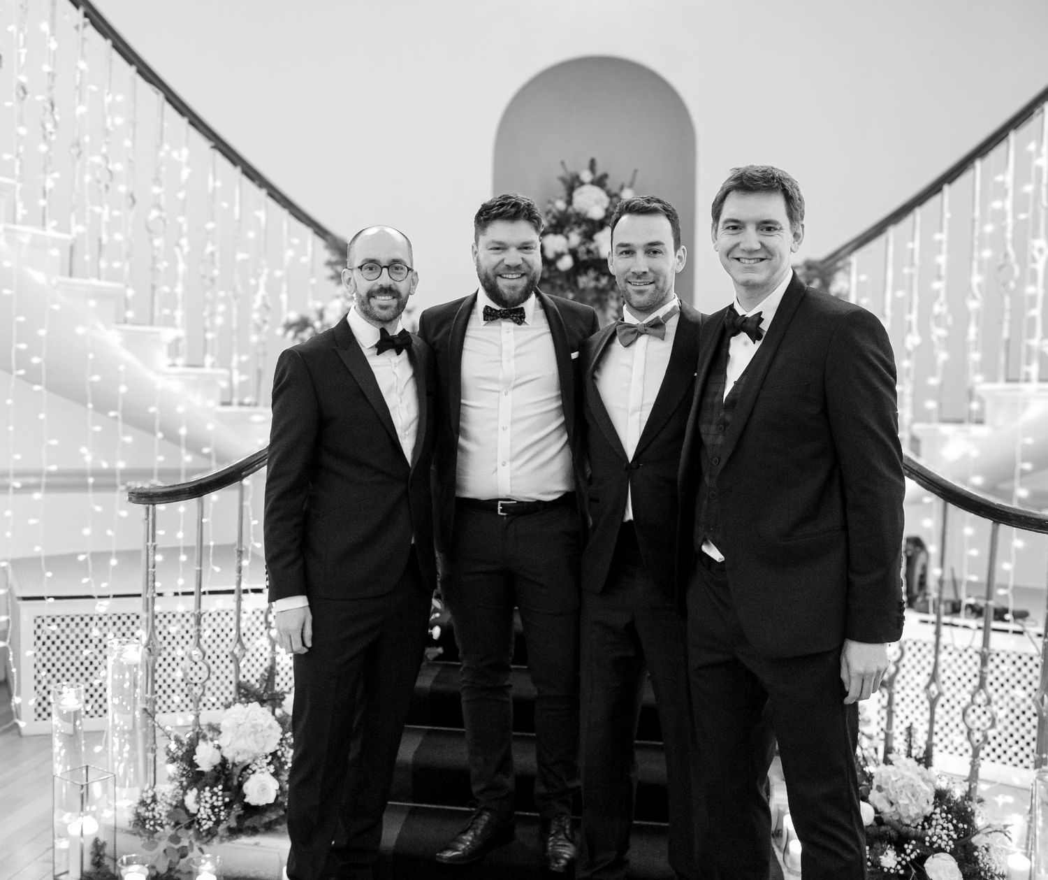 A black and white photo shows four men in formal tuxedos posing together at an elegant event setting.
