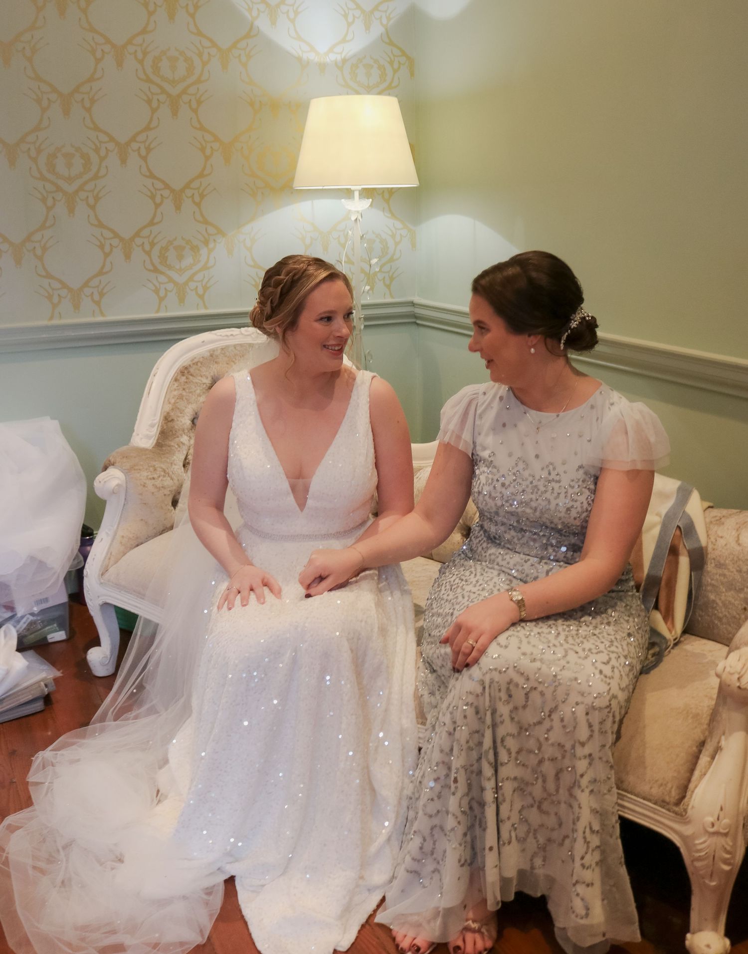 Two people in elegant dresses share a joyful moment seated on an antique chair with decorative wallpaper in the background.