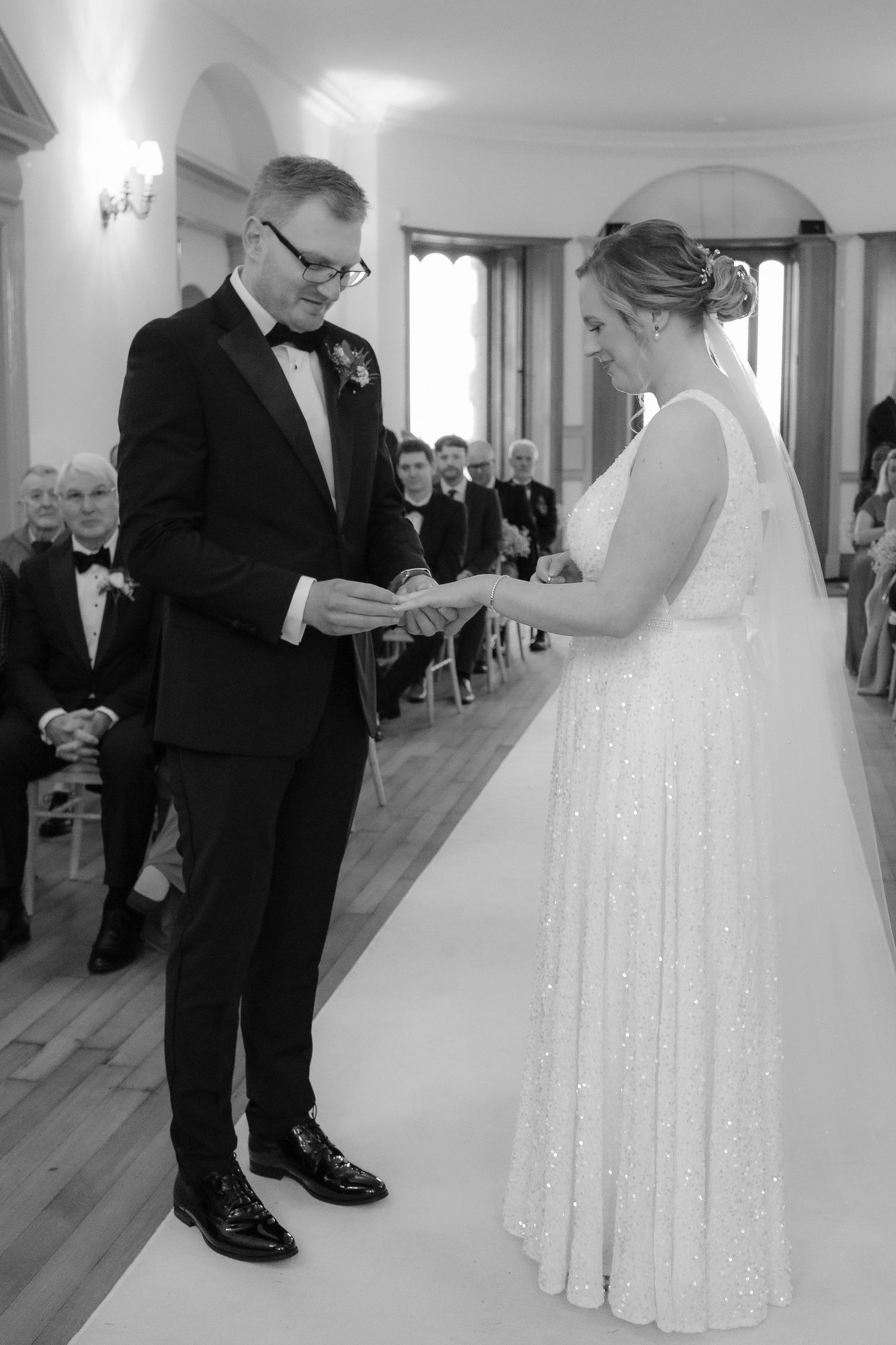 A wedding ceremony in a bright indoor venue shows two people exchanging rings during a formal service.