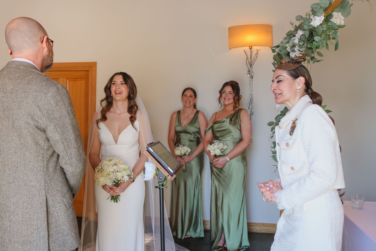 A bride in a white dress stands with their wedding party in sage green dresses during an indoor ceremony.