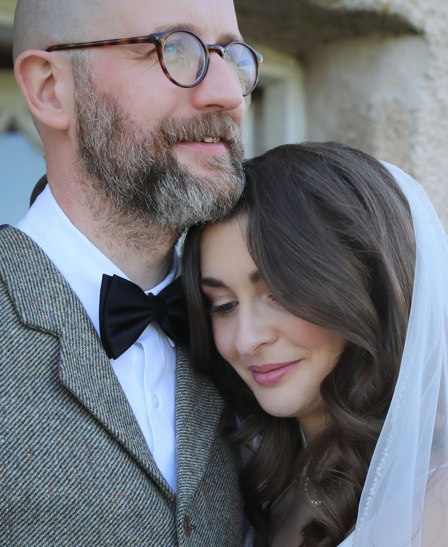 Newlywed couple in intimate wedding photo showing groom in tweed and bowtie with bride wearing white veil.