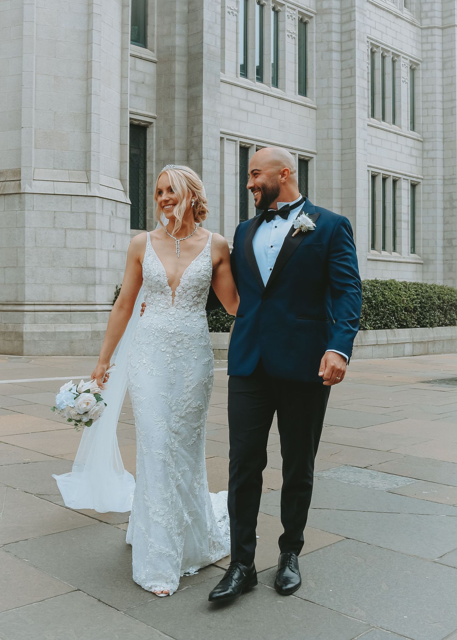 A newlywed couple walks together outside a grand stone building, with the bride wearing a lace gown and groom in navy suit.