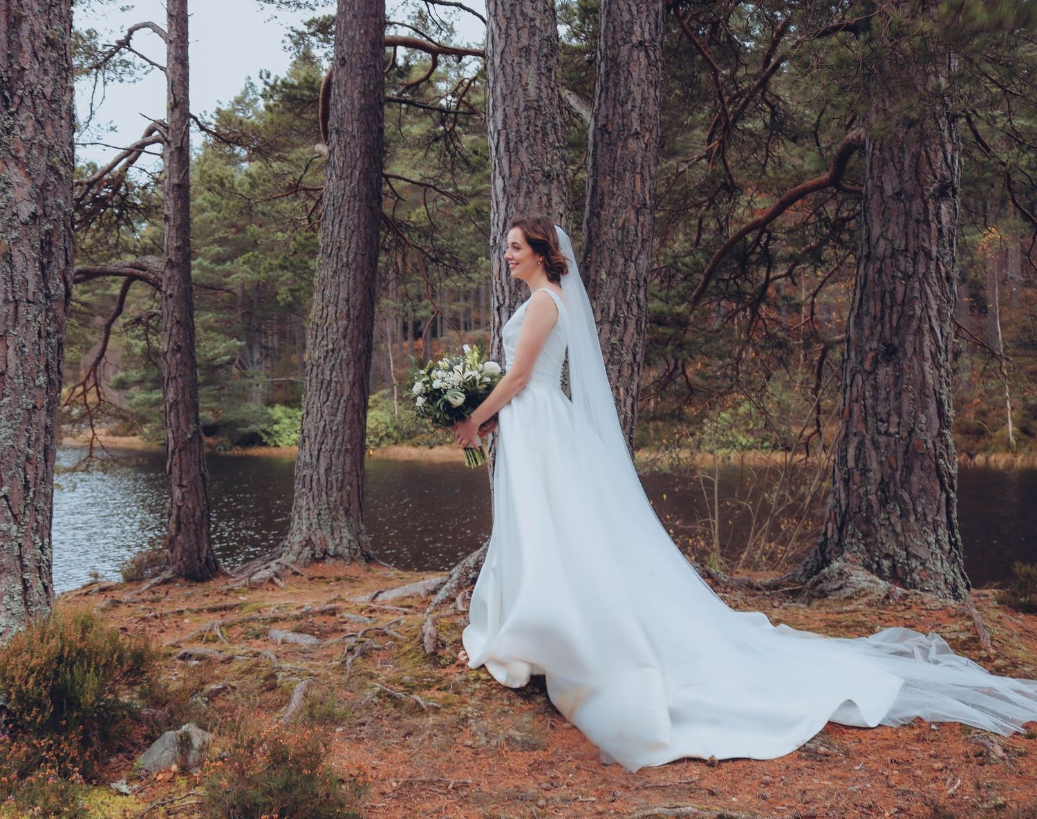 A person in a flowing white wedding dress with a bouquet stands on pine-covered lakeside cliff during autumn.
