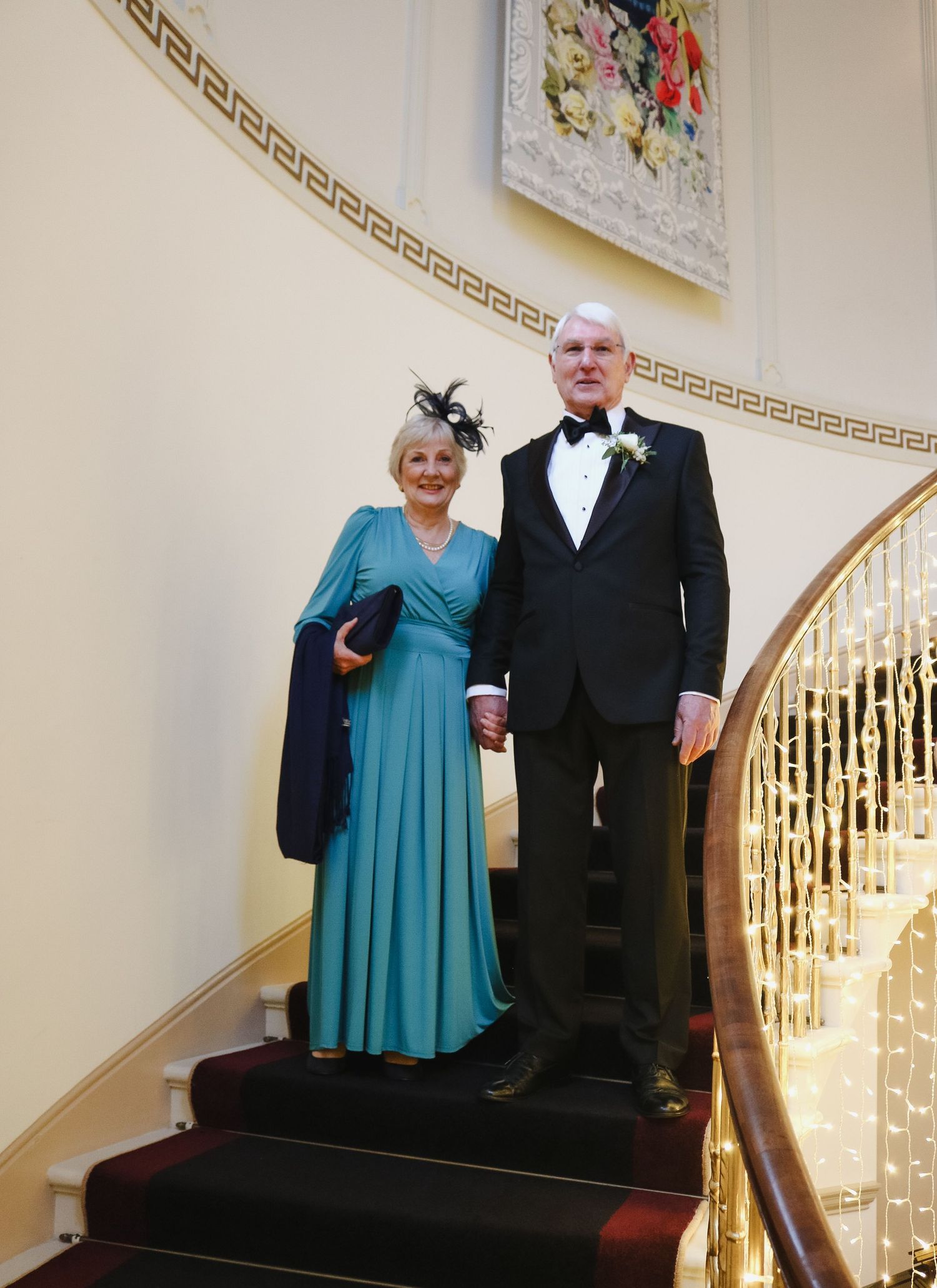 A couple in formal attire stands on an elegant curved staircase with gold railings and red carpeting.