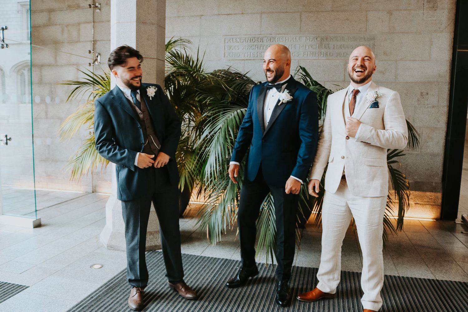 Three groomsmen in formal attire share a candid moment together by a tropical plant against a light wall.