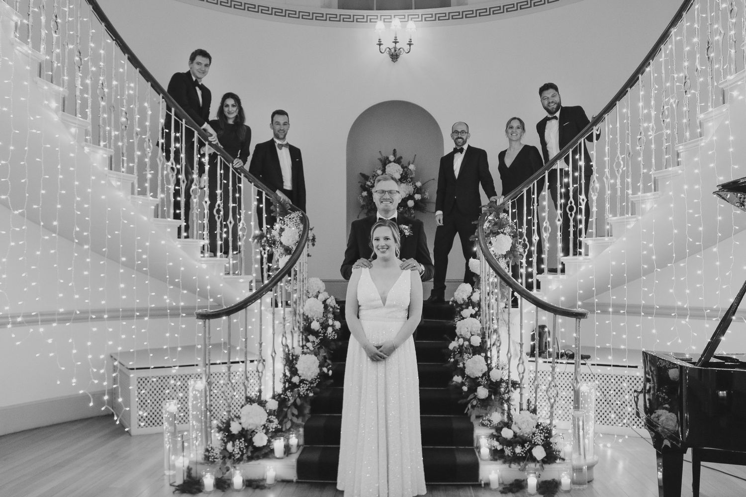 Black and white photo of a curved double staircase decorated with holiday lights and flowers at an elegant venue.