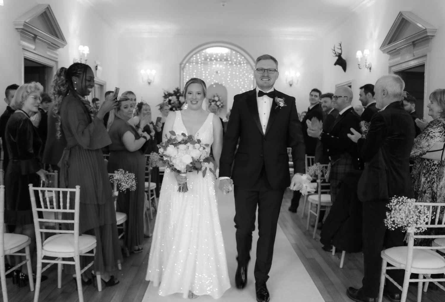 A couple walks down the wedding aisle in a historic venue while guests stand and capture the moment in black and white.