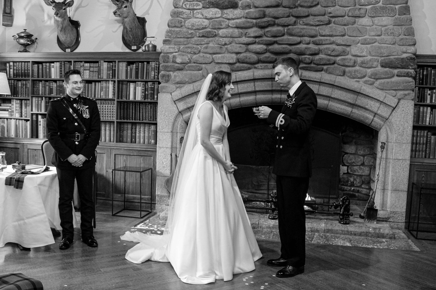 A wedding ceremony takes place in front of a stone fireplace with bookshelves and mounted deer heads in the background.