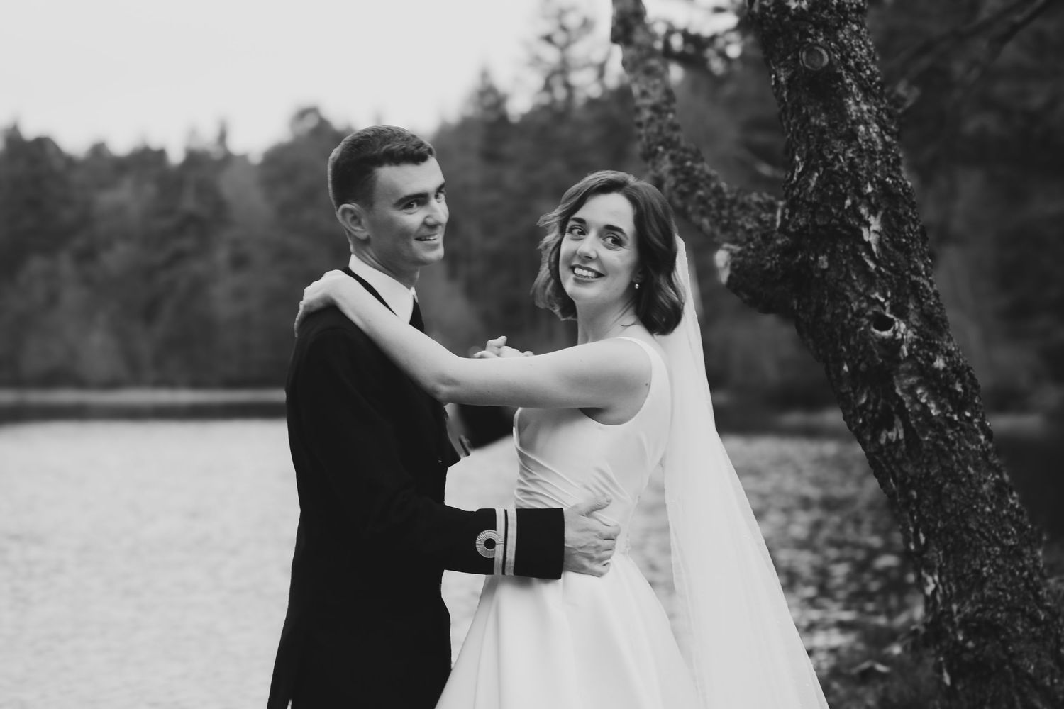 A black and white wedding photo of a couple dancing together by a lake with trees in the background.