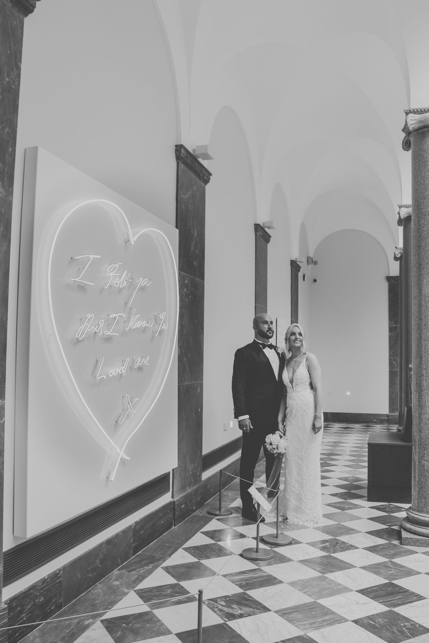 Black and white photo of a wedding couple standing in an ornate hallway with arched ceilings and checkered floor.