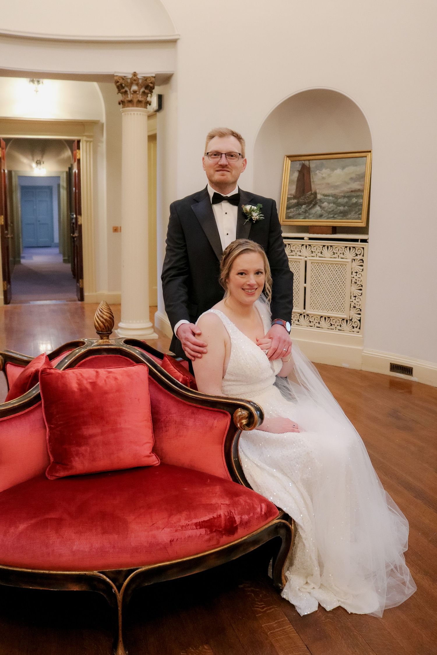 A wedding couple poses on an elegant red velvet chaise lounge in a richly decorated interior hallway.