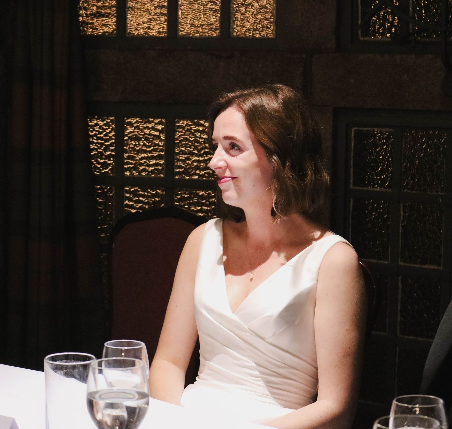 A woman in a white sleeveless dress sits at a dining table with water glasses, smiling towards the window.