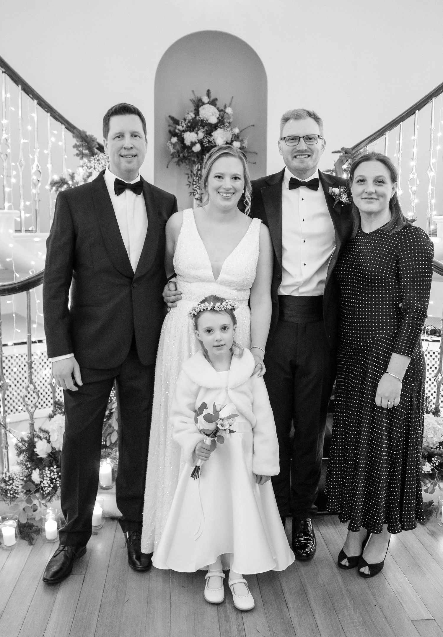A formal black and white wedding photo features a small group in evening wear and a flower girl at the staircase.