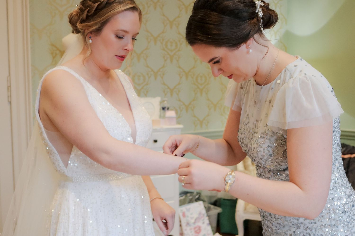 Two people in elegant white dresses help each other with final preparations in a room with patterned wallpaper.