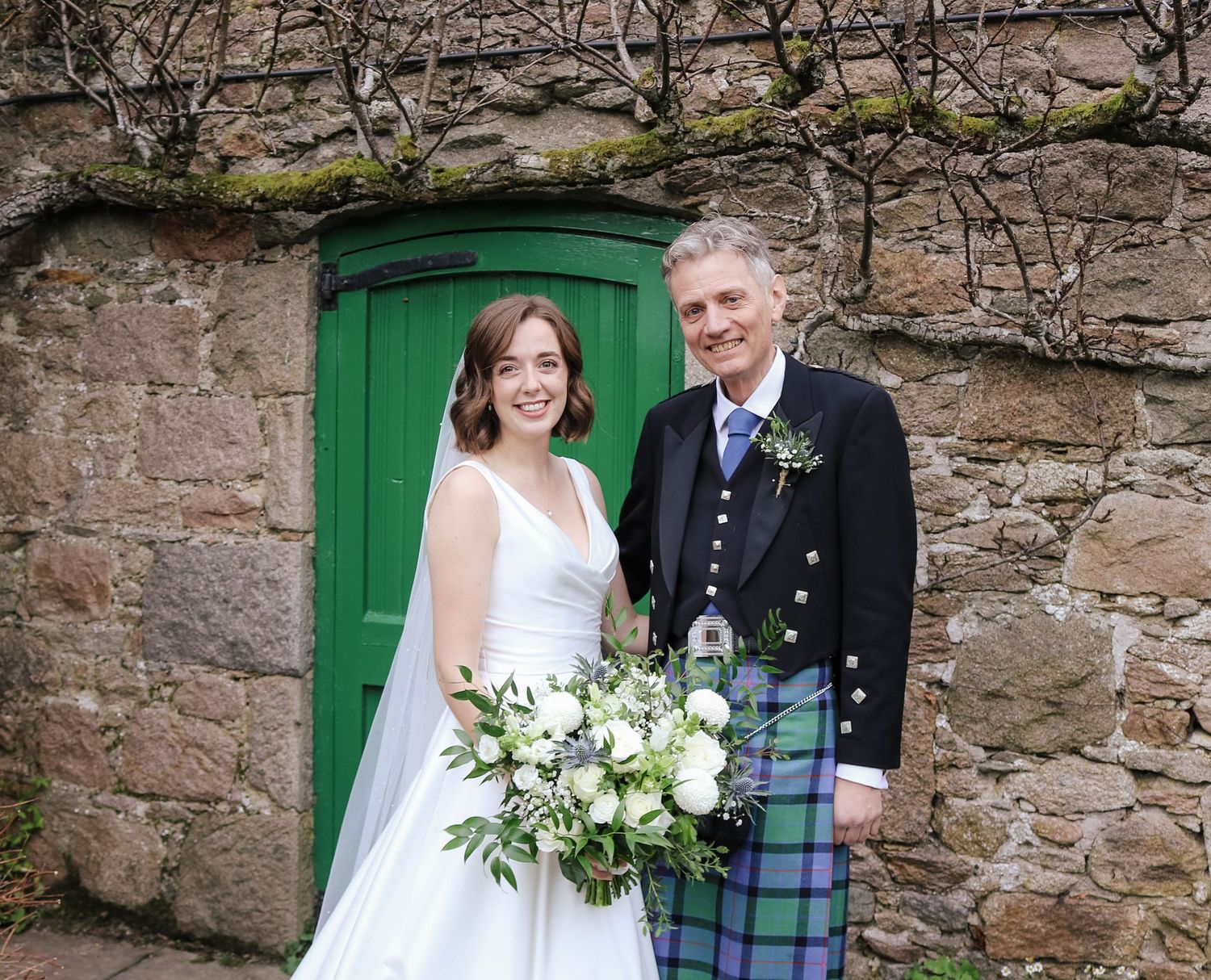 A bride and groom stand in front of a green door, with stone walls and vines, in Scottish wedding attire.