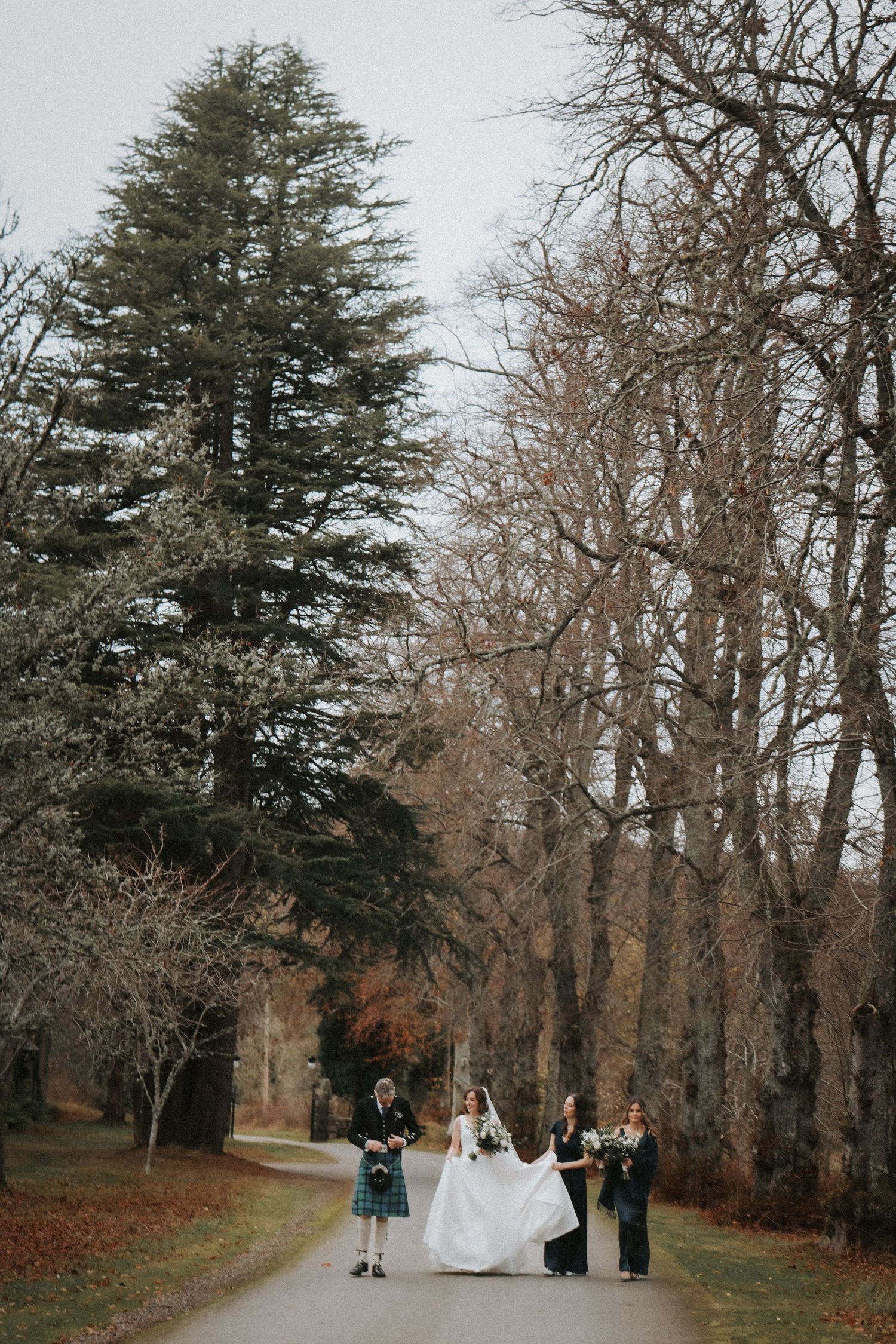 Wedding party poses along a scenic tree-lined path on an autumn day amidst fallen leaves and bare branches.