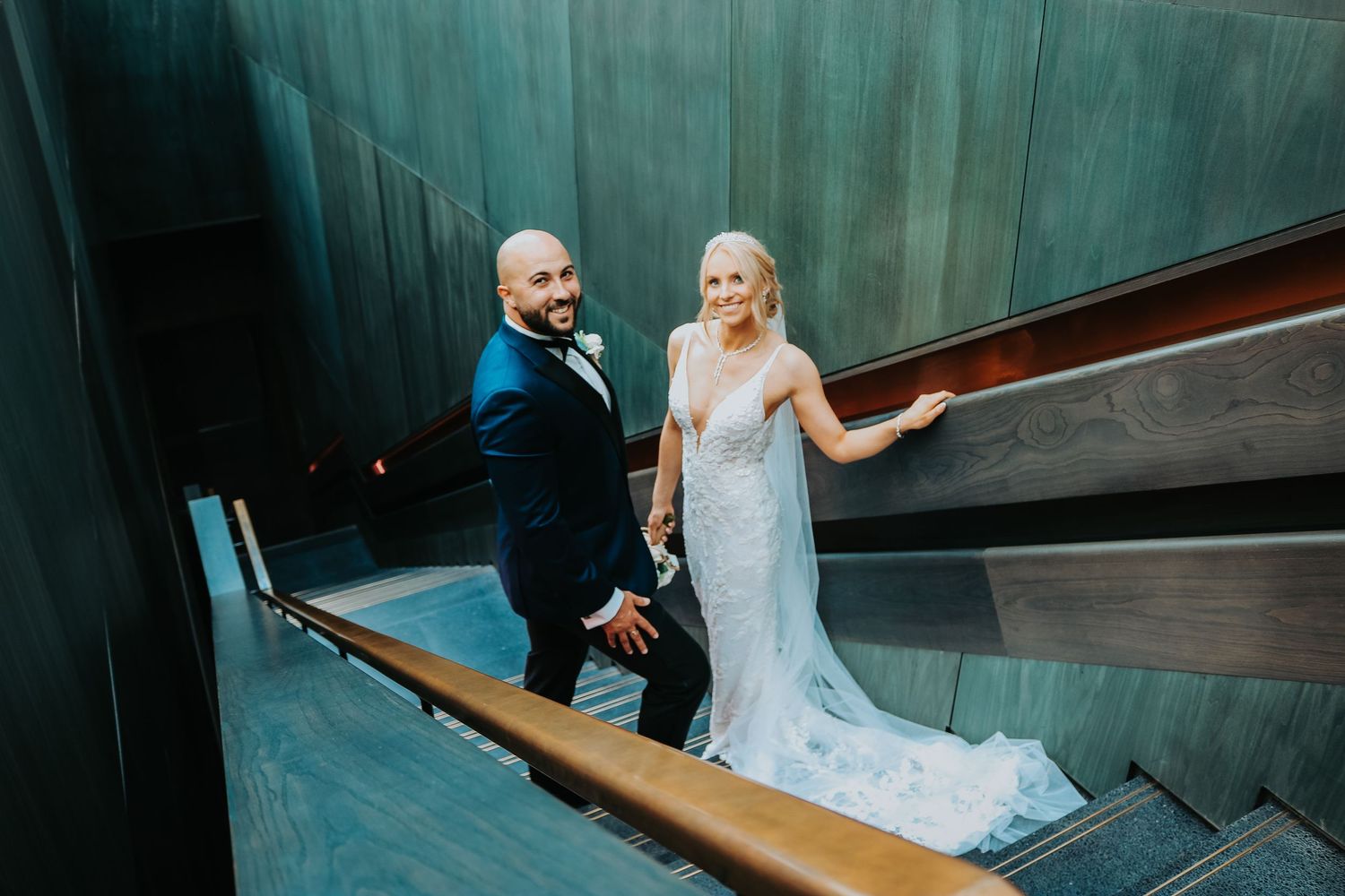 A couple in wedding attire ascends modern geometric stairs with teal-colored walls in an artistic venue.