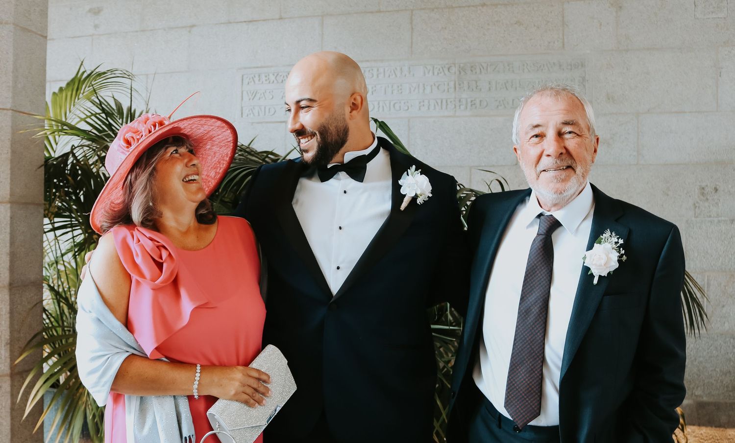 Wedding guests wearing formal attire including a pink sun hat, black tuxedo and navy suit gather together outdoors.