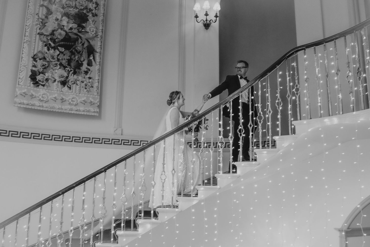 Two figures descending an elegant white staircase with ornate railings in a mansion or grand venue in black and white.