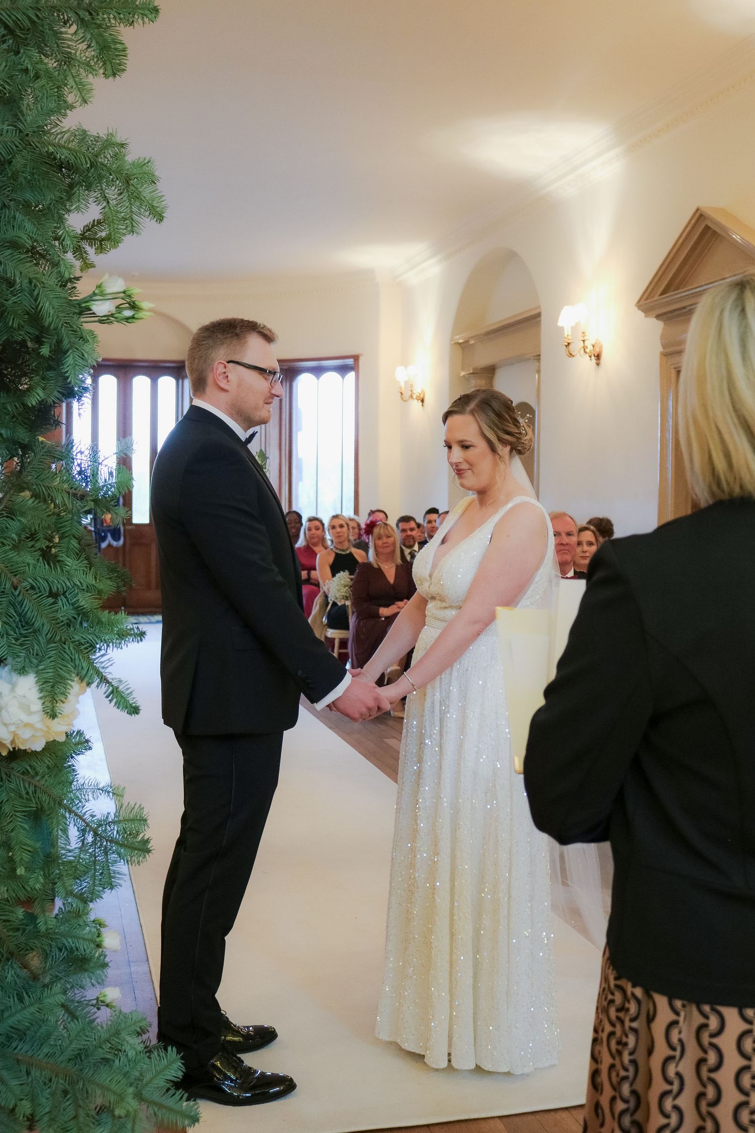 Wedding ceremony in a bright indoor chapel with a large Christmas tree and wall sconces illuminating the elegant space.