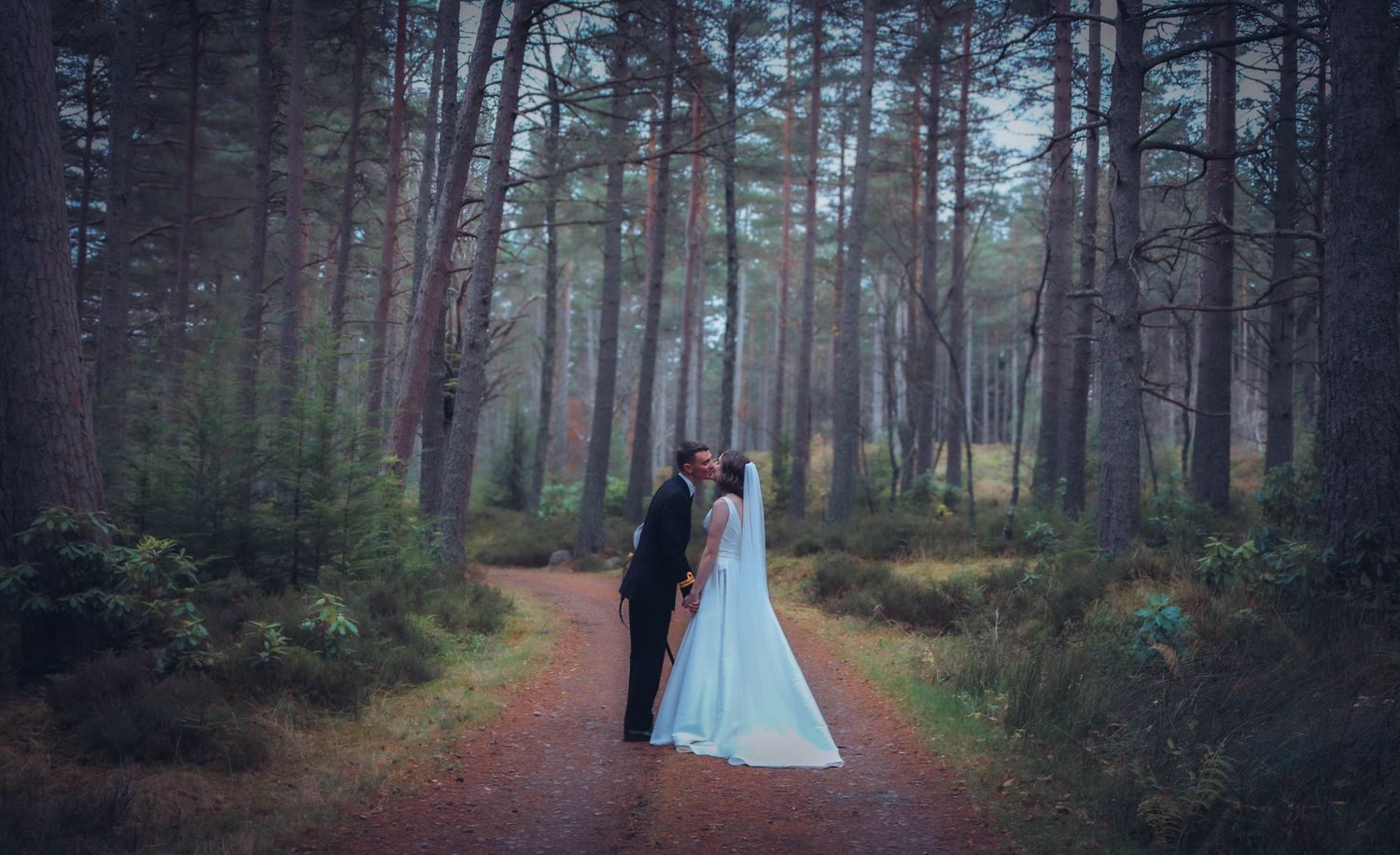 A couple stands on a misty forest path wearing formal attire with a flowing blue gown creating a magical woodland scene.
