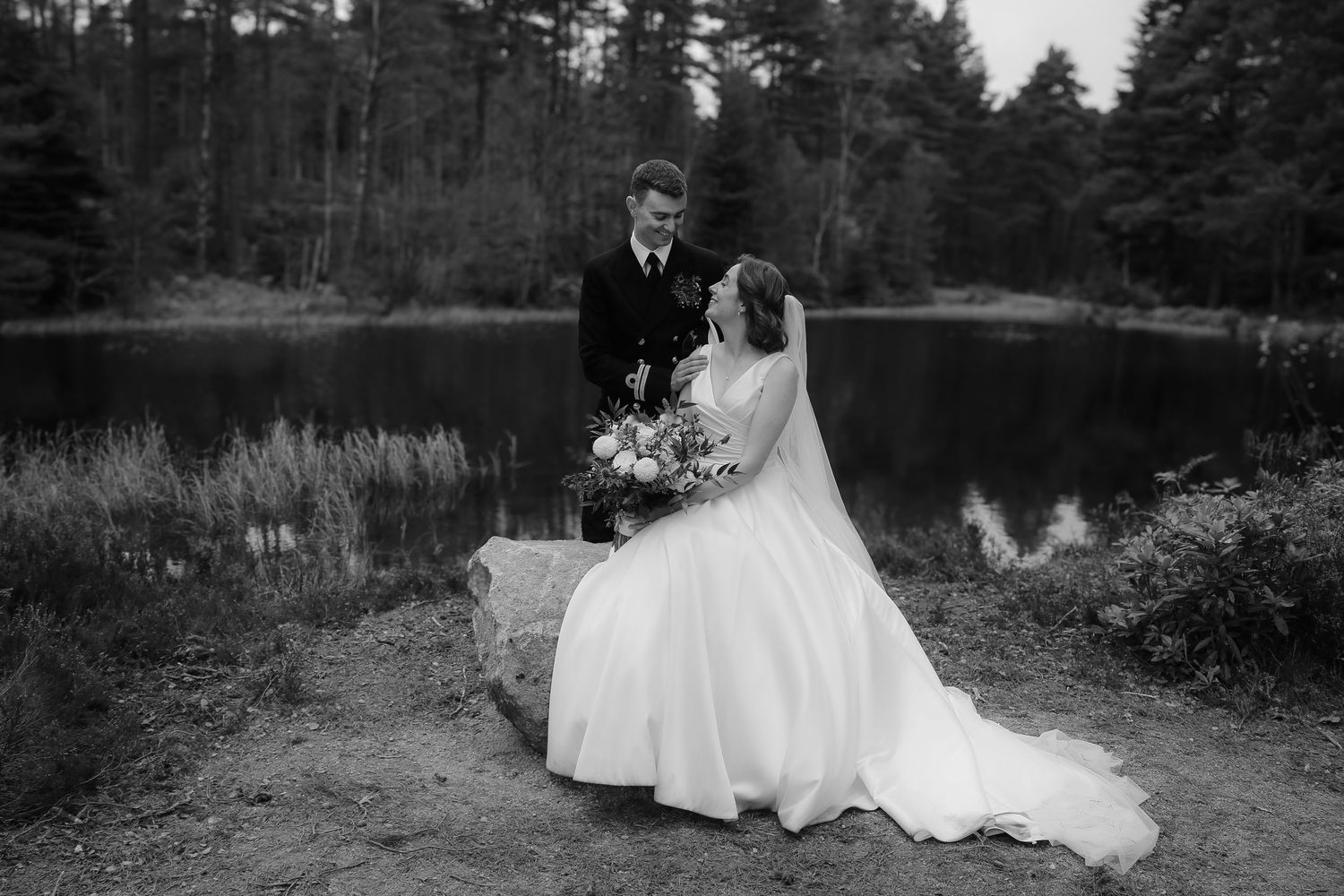 A black and white wedding photo of a couple standing by a serene lake surrounded by pine trees.