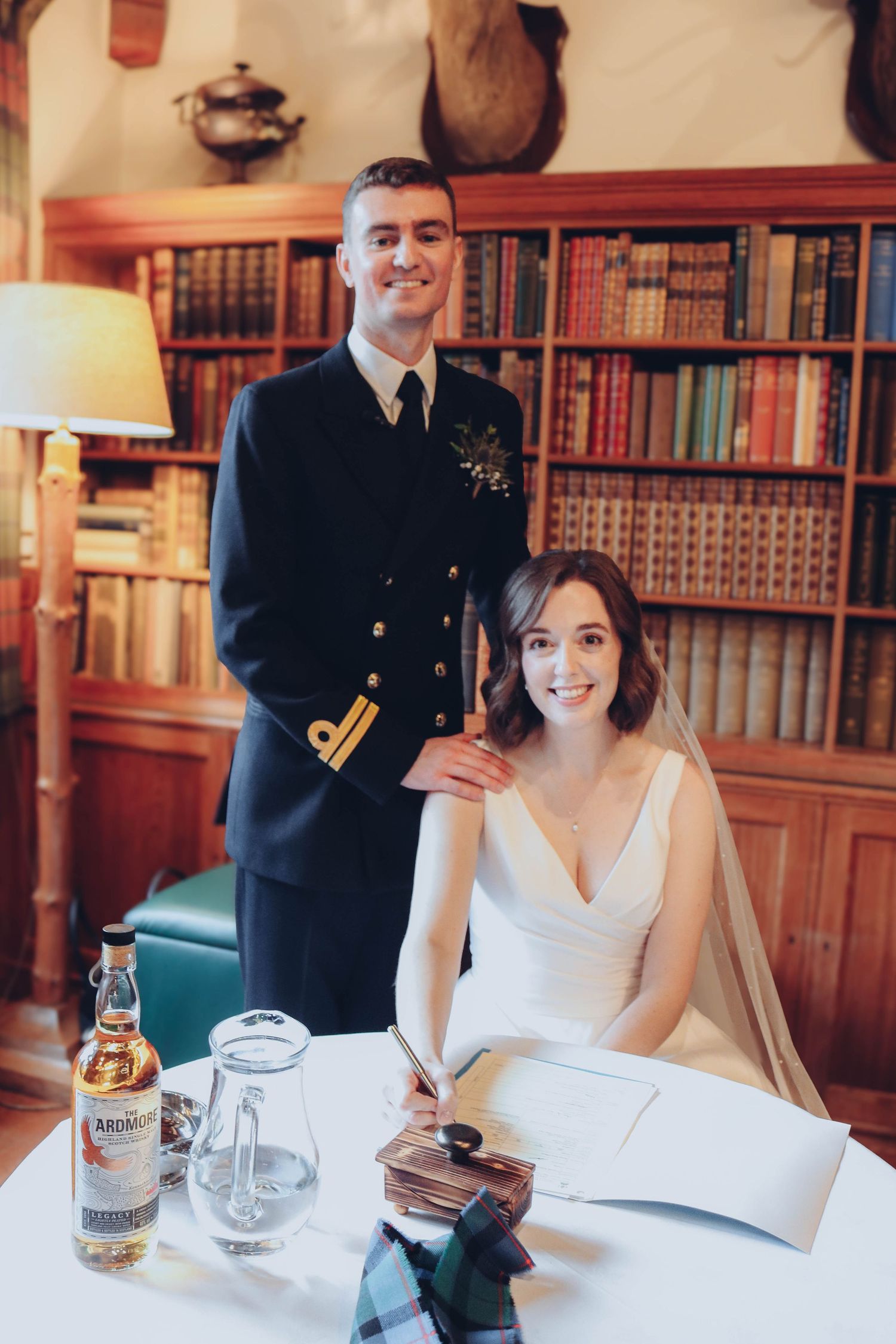 A bride in white dress and military groom pose in a study with bookshelves and a vintage lamp beside them.