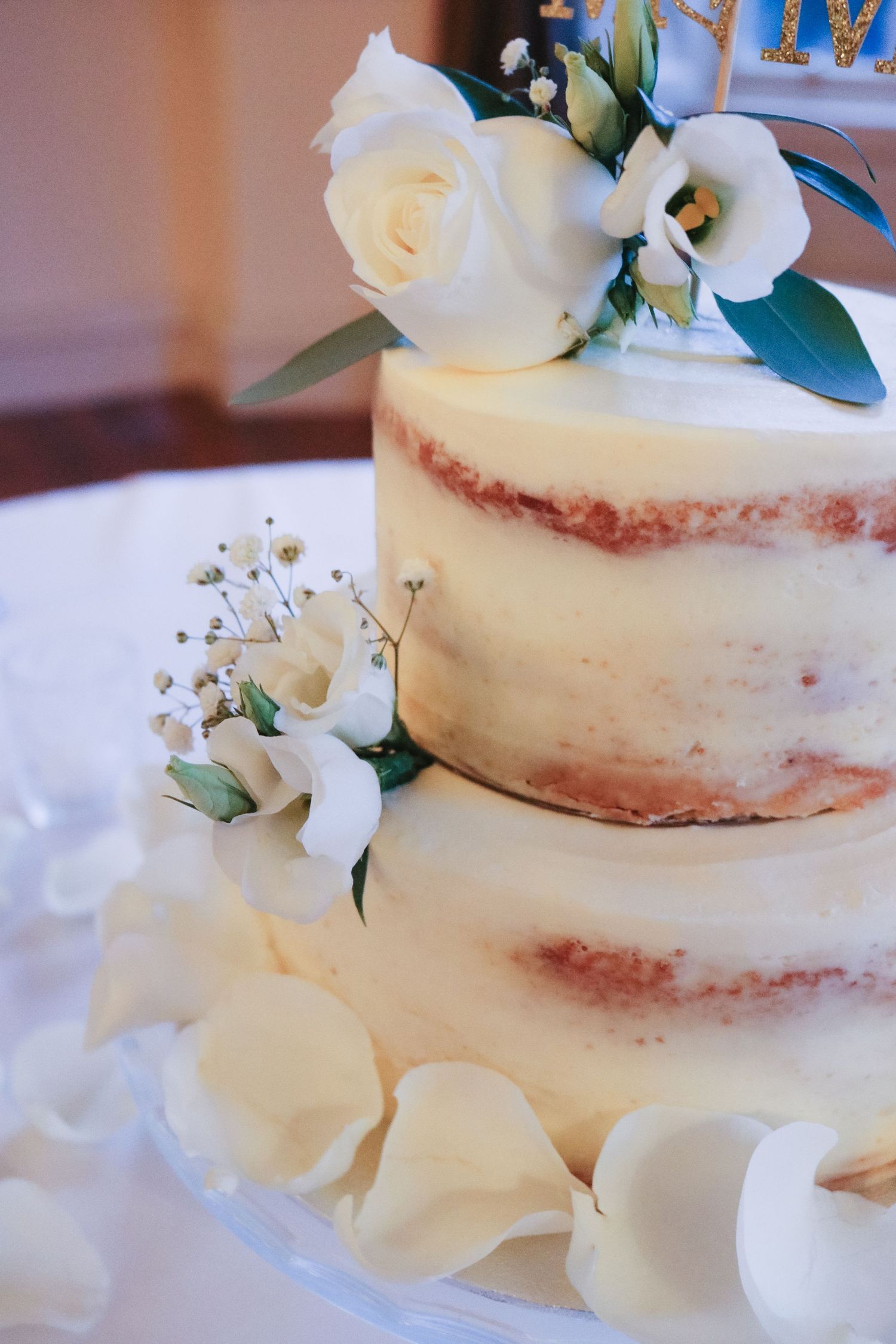 A semi-naked wedding cake decorated with white roses and greenery features a rustic, partially frosted design.