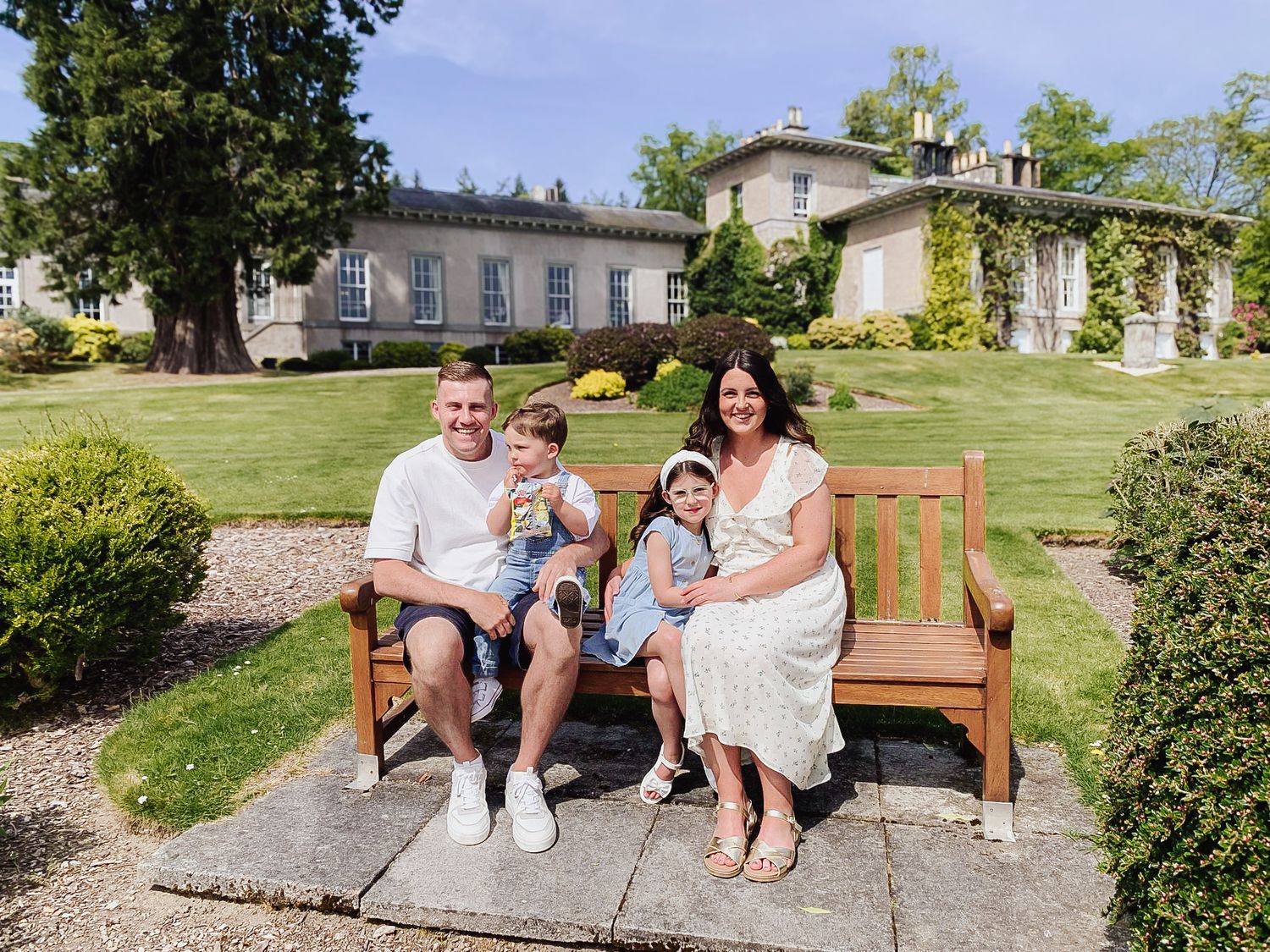 Family sitting on a wooden garden bench with landscaped grounds and elegant buildings in the background.