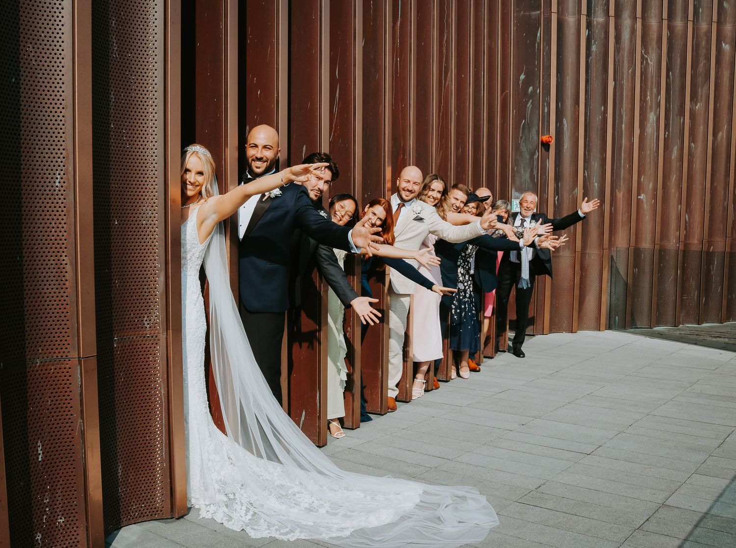 A wedding party poses creatively against a rustic metal wall during celebrations.