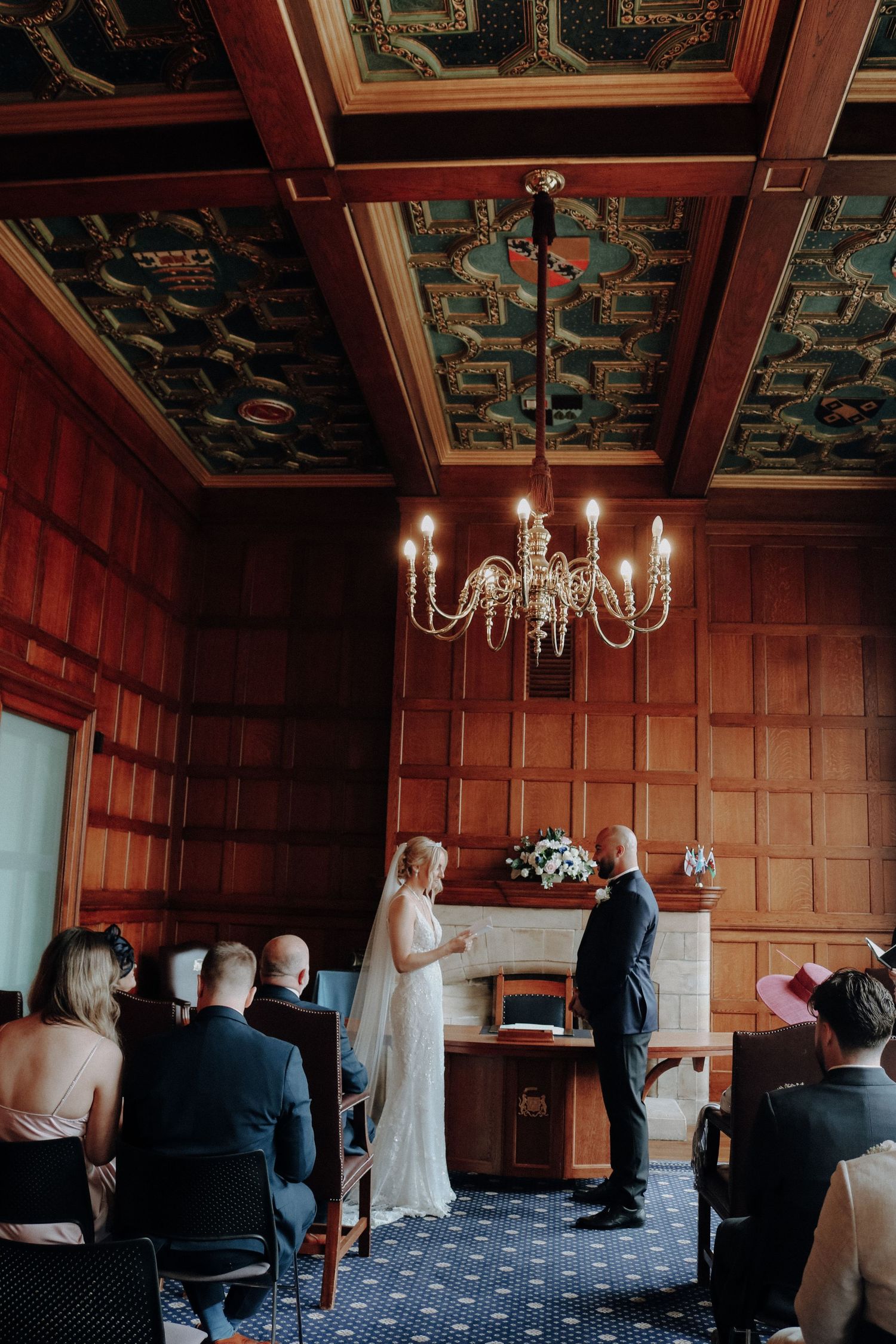 An intimate wedding ceremony in an elegant wood-paneled room with a crystal chandelier and ornate ceiling details.