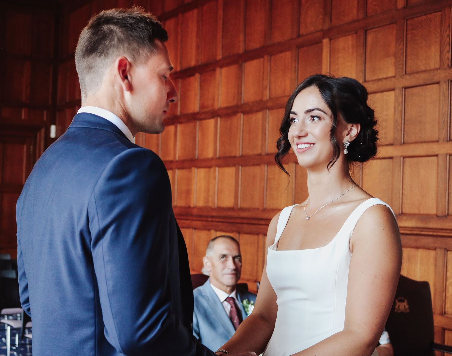 A bride and groom exchange vows in an elegant wood-paneled room while a suited guest looks on in the background.