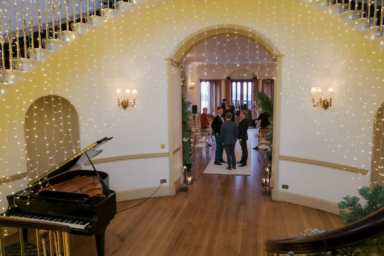 Elegant event space with hardwood floors, string lights, grand piano, and arched doorways creating a romantic atmosphere.