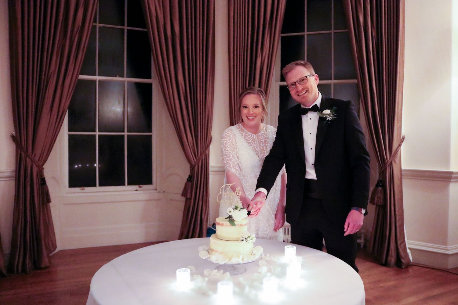 A couple in formal attire cuts their wedding cake together at an elegant reception with burgundy curtains and candlelight.