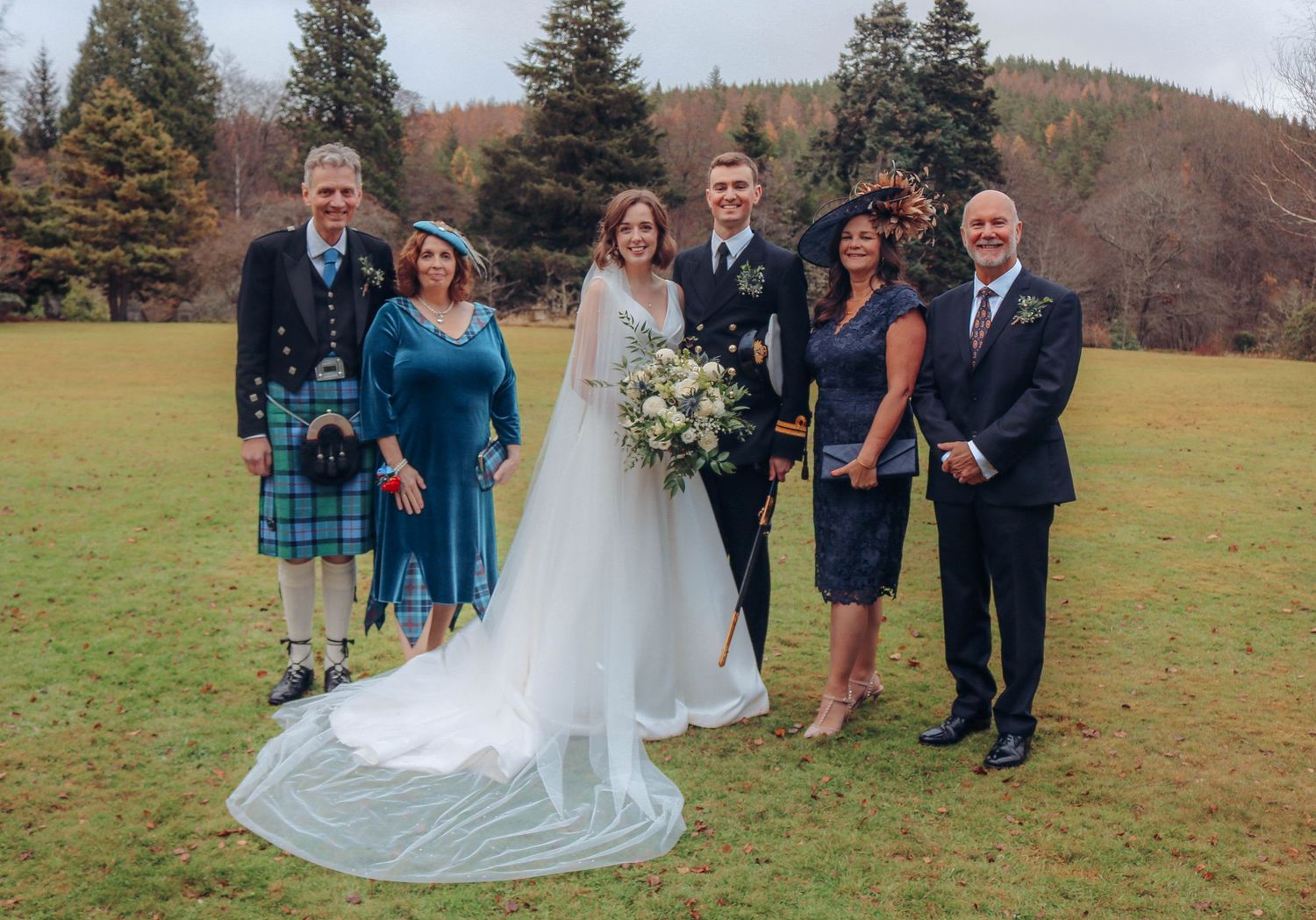 Wedding party stands together on a green lawn with autumn trees in the background.