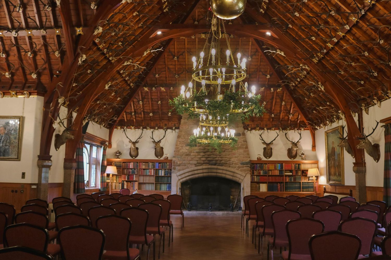 Large historic lodge room with exposed wooden beams, chandelier, stone fireplace and mounted deer heads on brick walls.