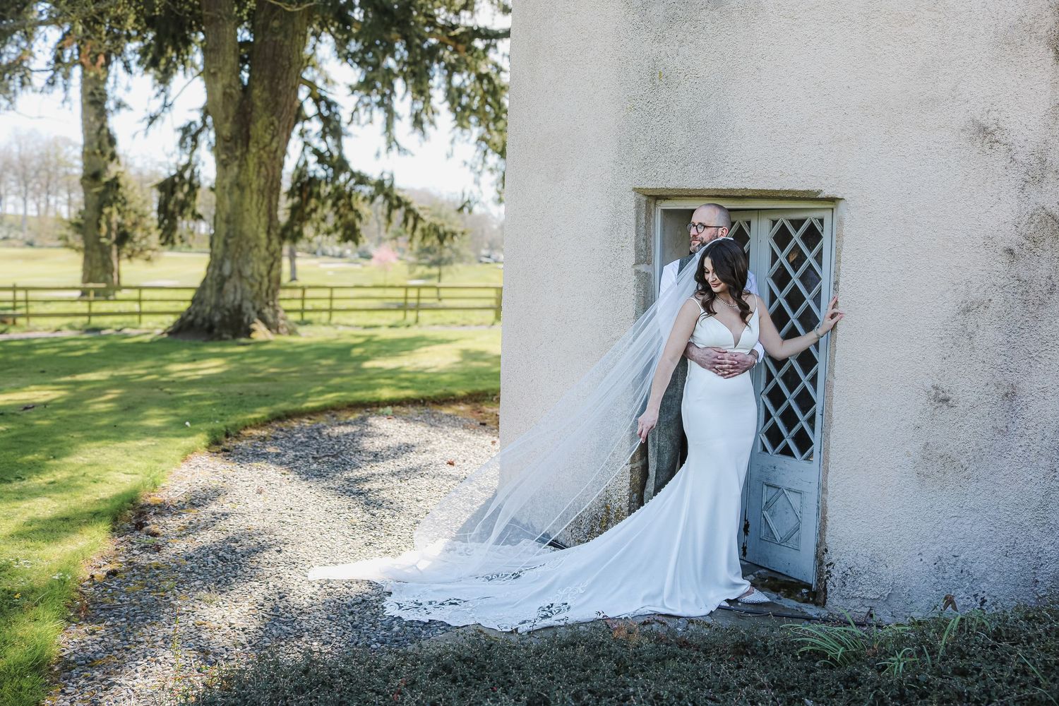A bride in a fitted white mermaid gown with long train poses elegantly by a rustic blue door at a countryside venue.