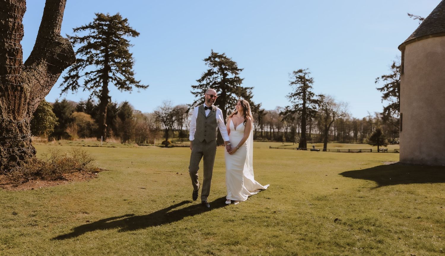 A couple walks together on a sunny day across a grassy lawn with tall pine trees in the background.