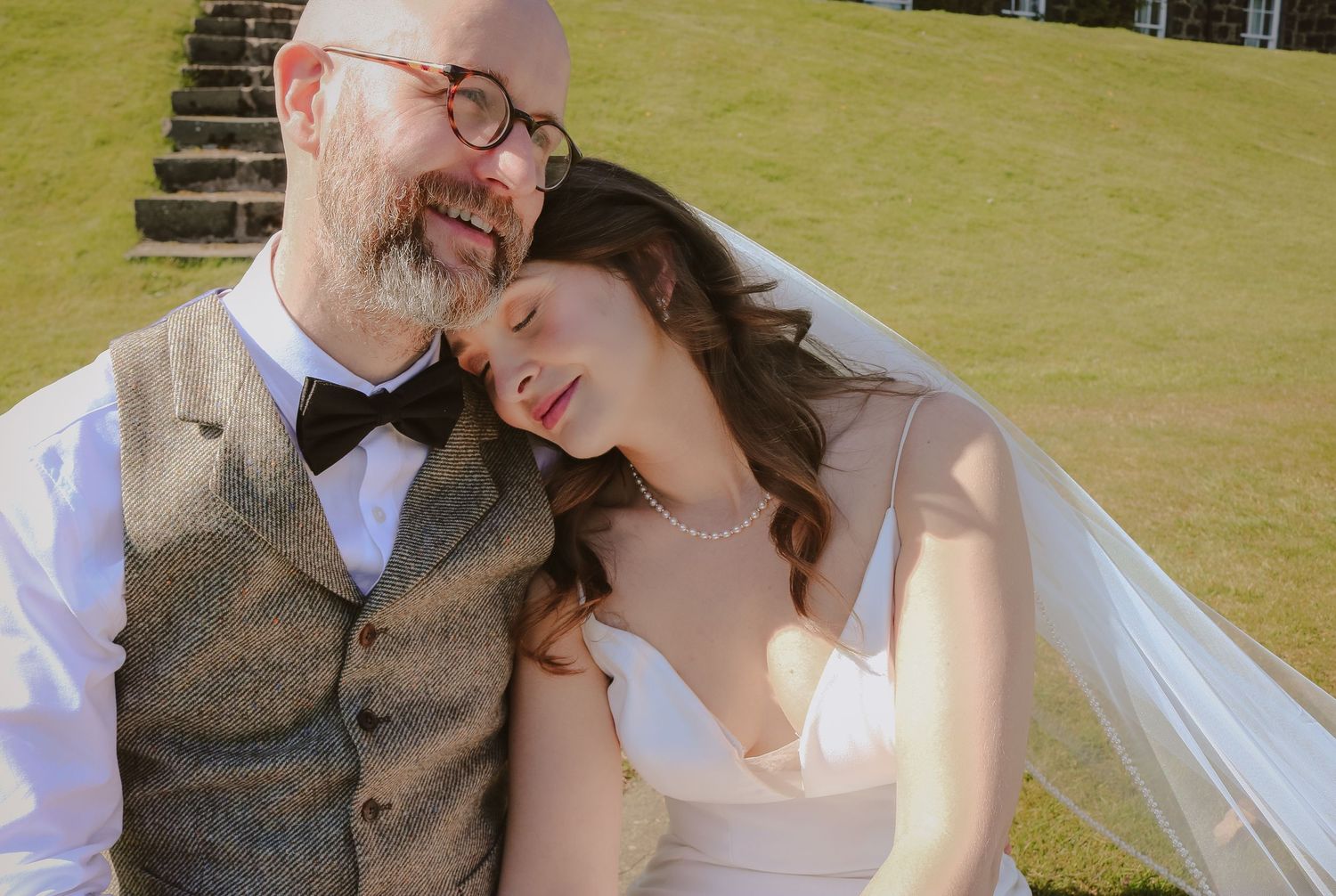 A happy couple shares an intimate moment outdoors on their wedding day, dressed in formal attire against a green lawn.