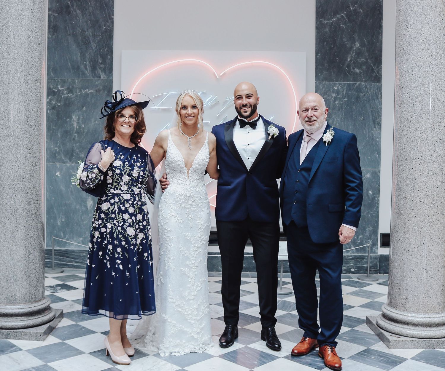 Wedding portrait with couple and family members in front of marble columns and pink neon heart.
