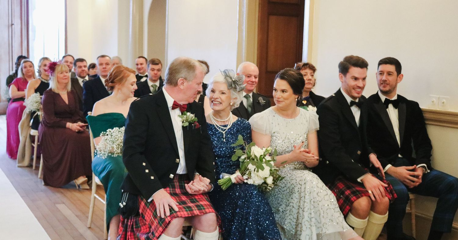 Wedding party sits in formal attire at a traditional church ceremony with attendees in colorful dresses and kilts.
