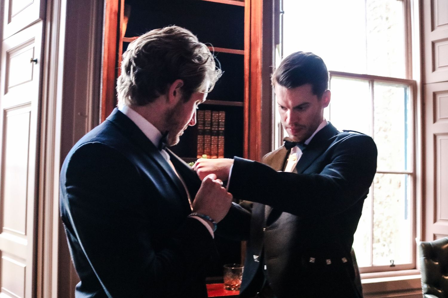 Two men in dark suits examine and discuss a document near a wooden window frame in a formal setting.