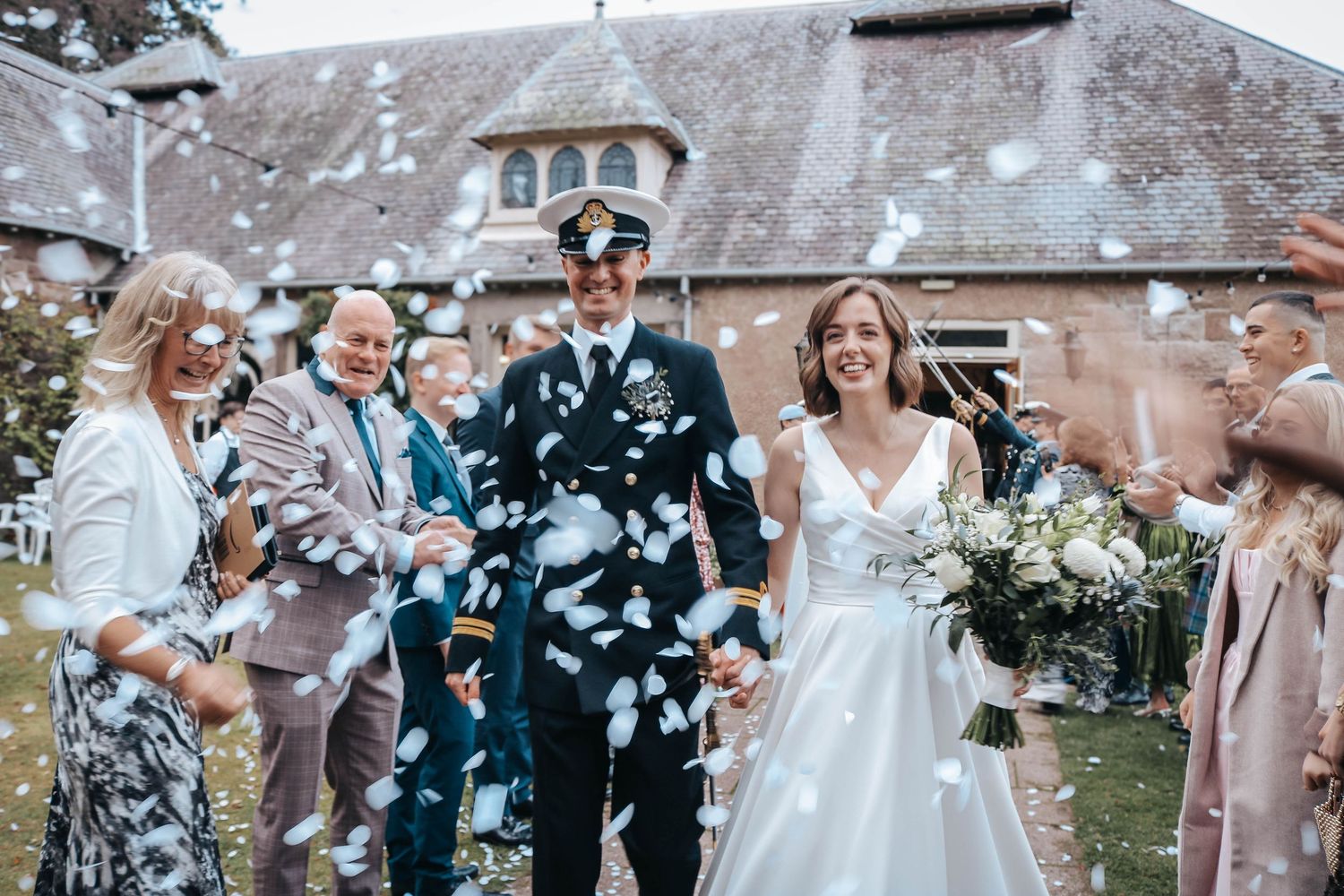Wedding guests shower newlyweds with white confetti during outdoor celebration at rustic church reception.
